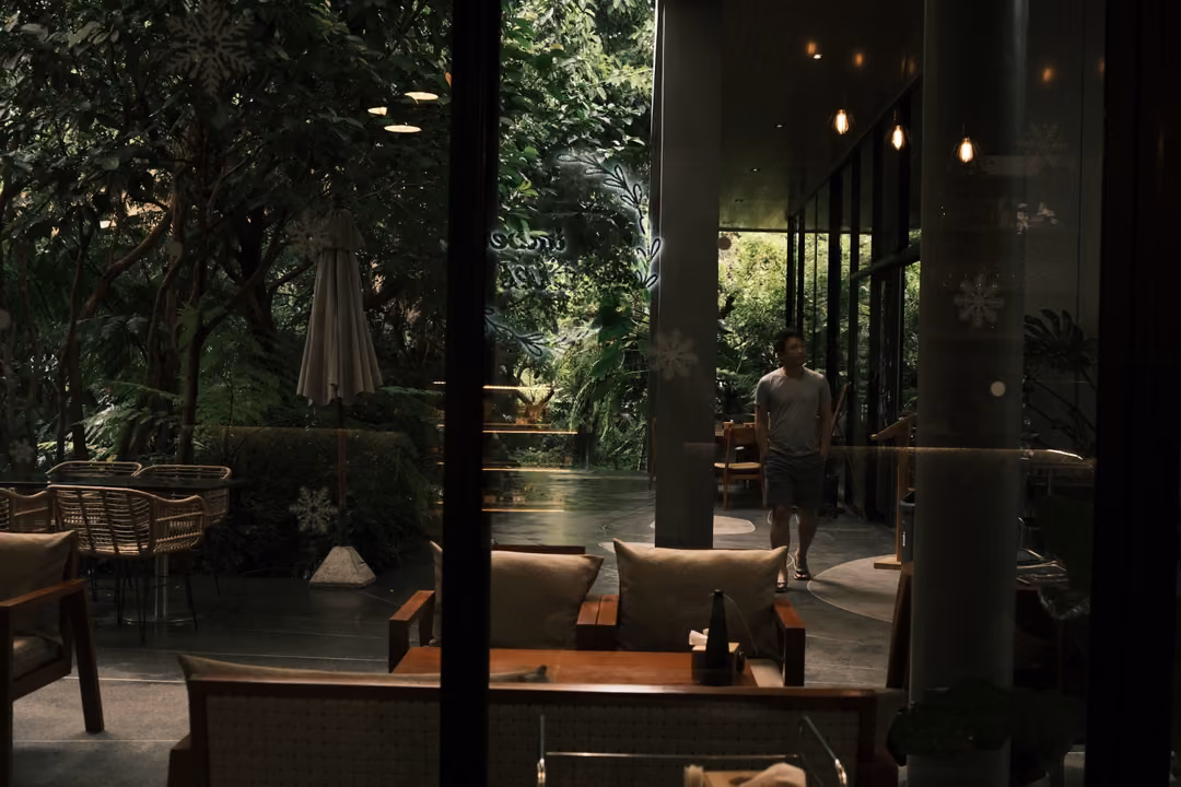 A dim café interior with pendant lights and wicker sofas, a garden and a walking figure seen through glass.