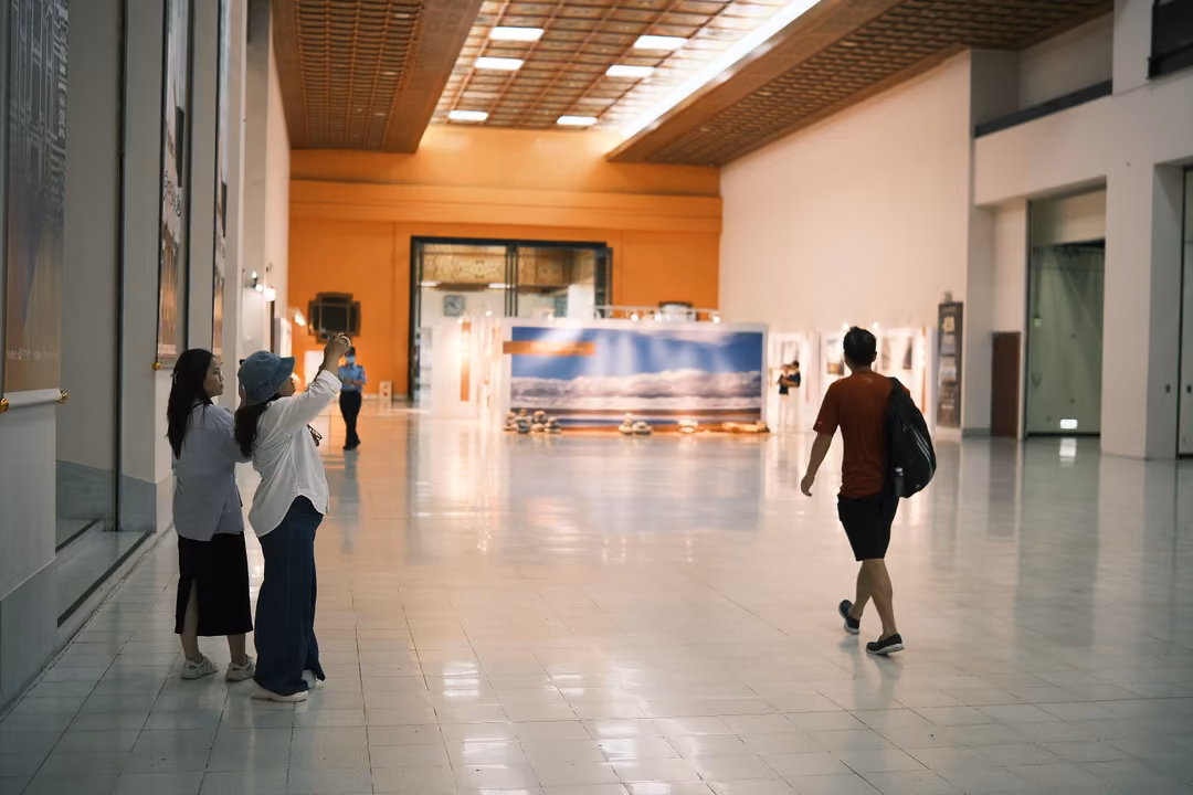 Long bright gallery with a polished floor, two visitors photographing a panel, another walking away with a backpack