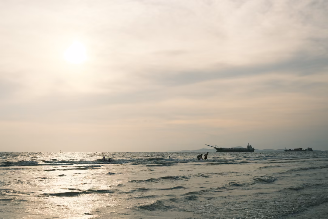 A hazy sun over a calm sea, a ship and a few small figures wading near the horizon