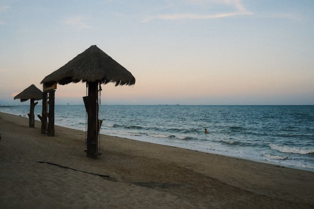 Two thatched beach umbrellas on wooden poles silhouetted on the sand at dusk, the sea and a lone swimmer