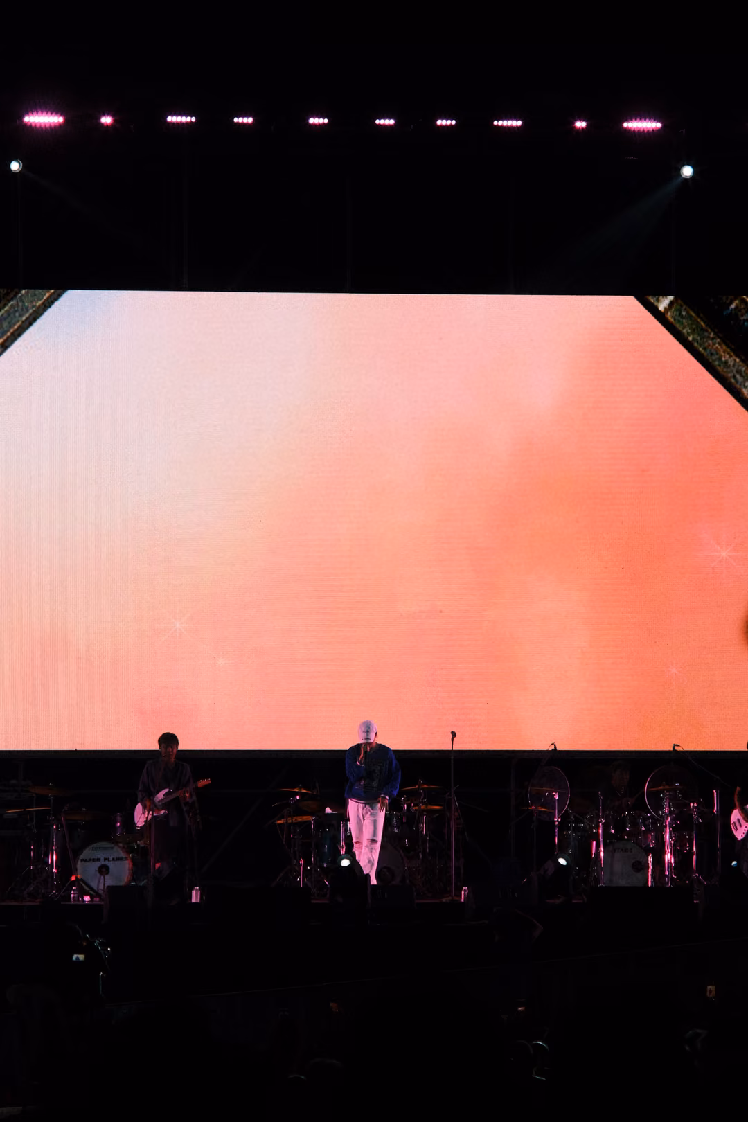 Vertical view of a soft pink and peach screen above the dark stage where a singer and guitarist perform.