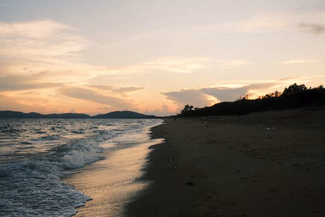 Empty beach at sunset, waves washing in, dark trees on a low point and peach clouds over distant hills