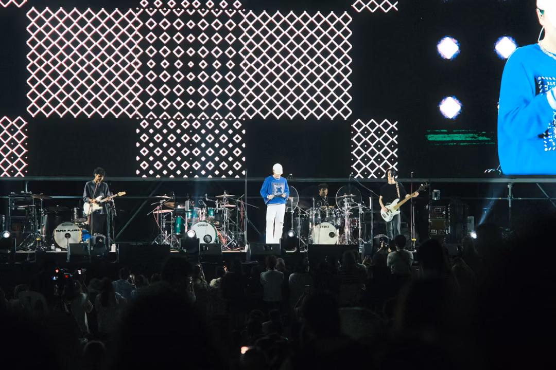 Band on stage under diamond-pattern screen panels, a close-up of the singer's blue sweatshirt at the right edge.