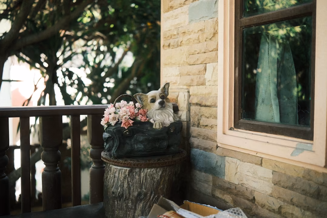 Dog figurine and pink artificial flowers on a stump planter beside a stone wall and window on a porch