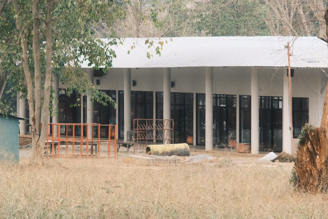 Low white building with tall columns and a metal roof behind a dry grass field and leaning trees