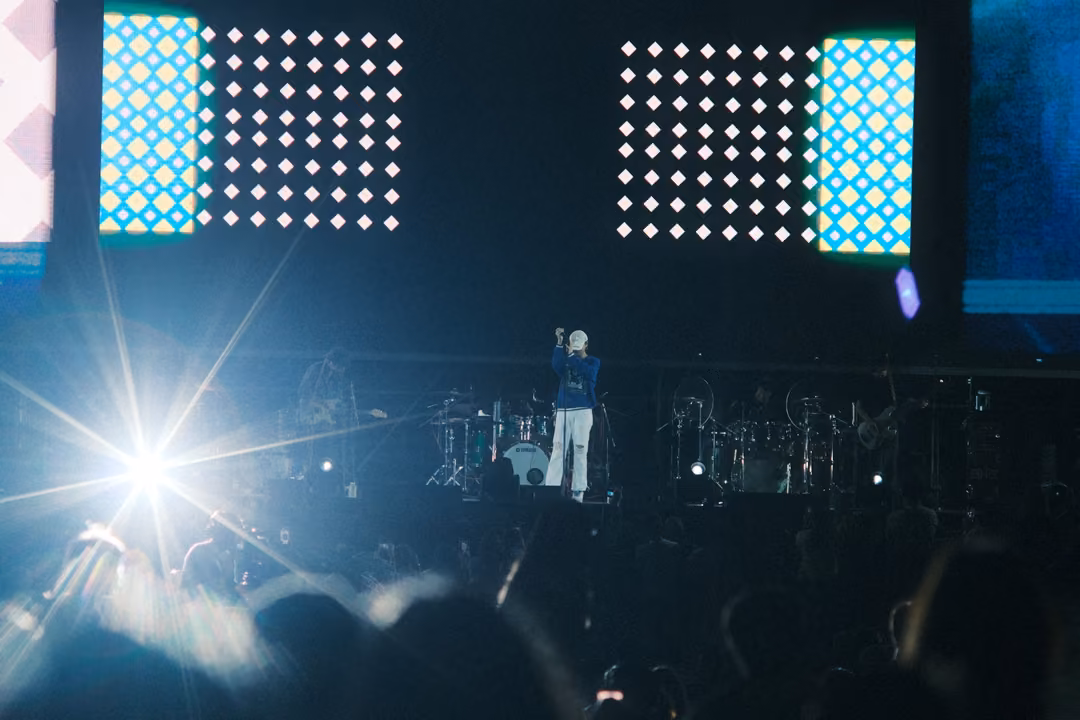 Stage with a bright starburst light at left, the singer at centre, screen panels with dotted grids and teal blocks.