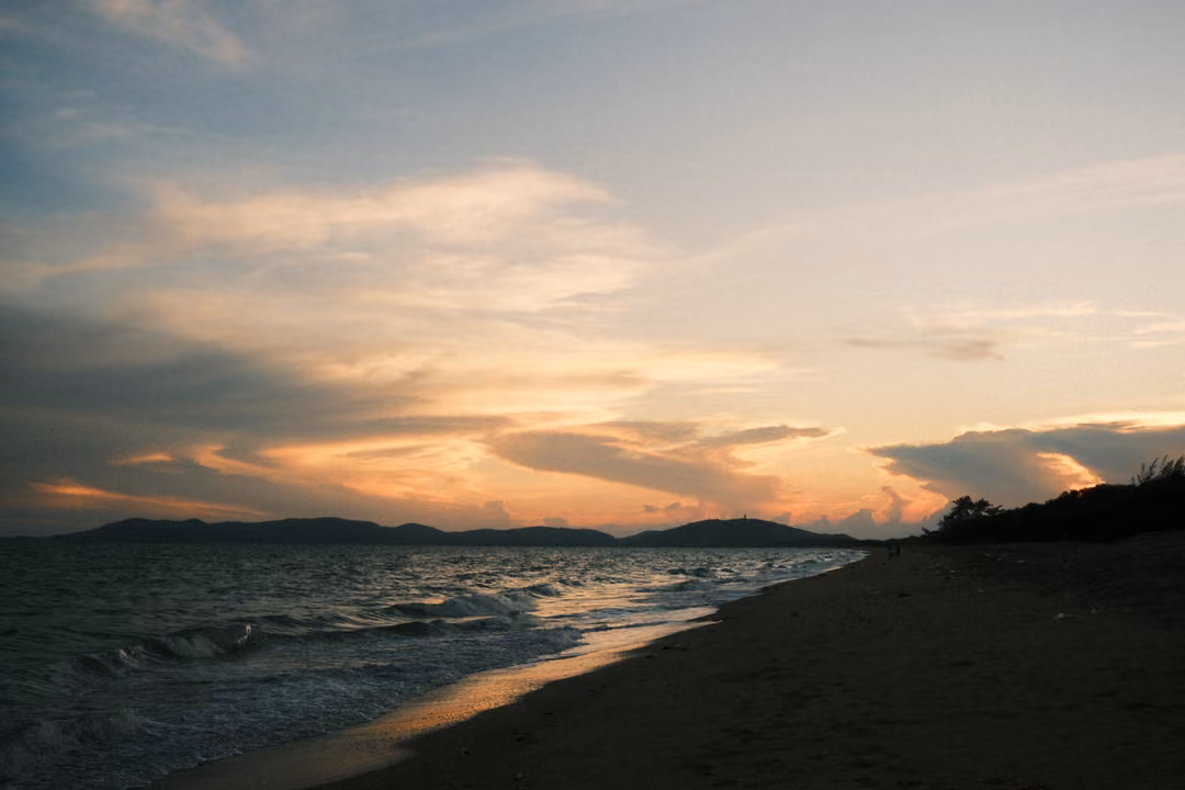 Sunset over a bay with streaked orange and grey clouds, dark hills, a curving shoreline and waves