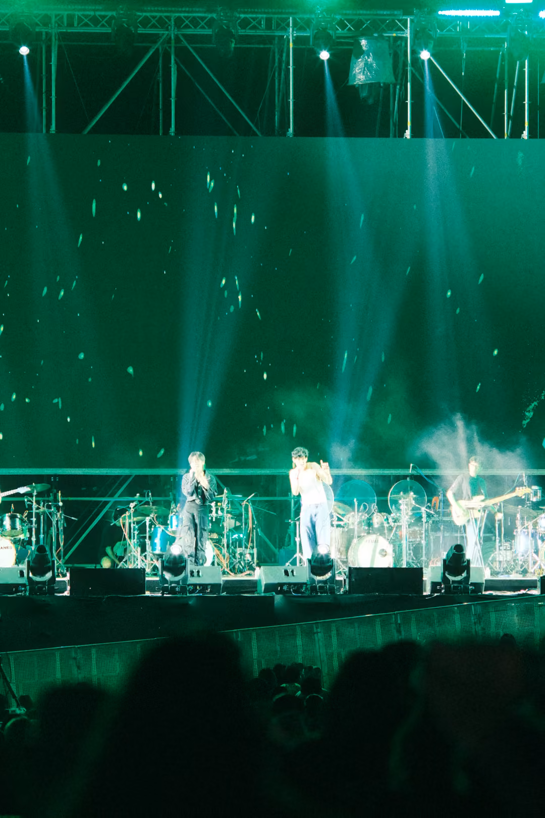 Vertical view of the stage in green light with smoke jets and spotlight beams, singer and guitarists performing.