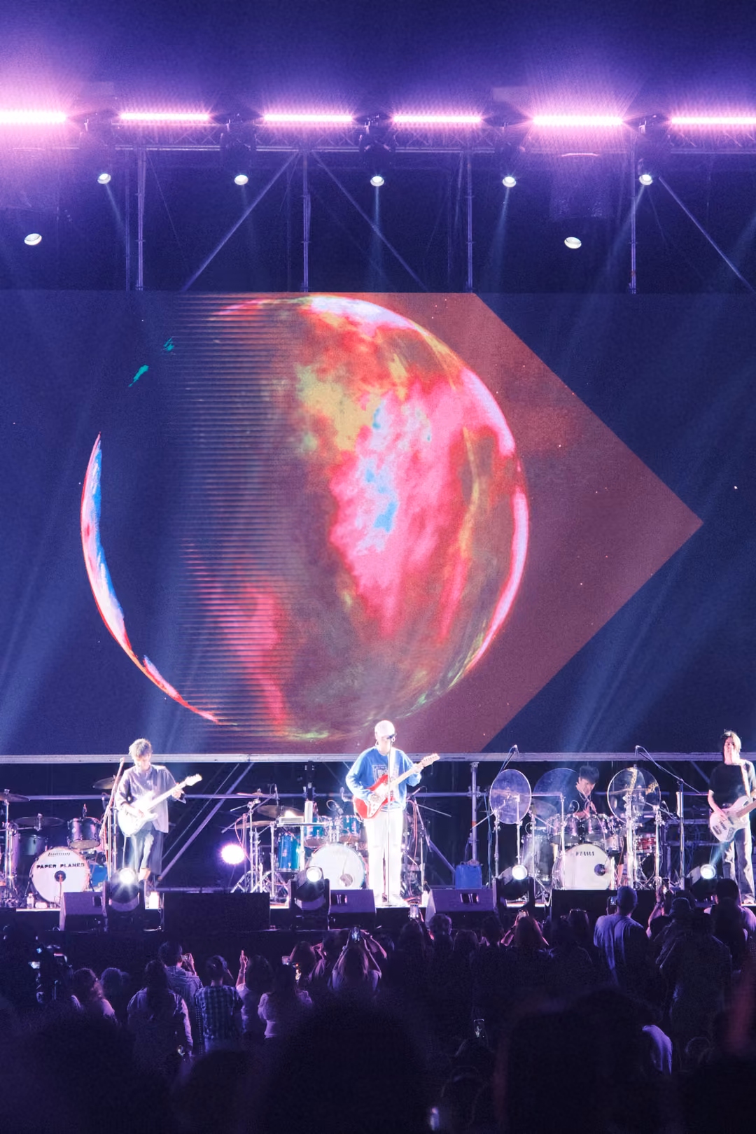 Vertical view of a stage with a glowing red sphere on screen, a singer with a red guitar and bandmates below.