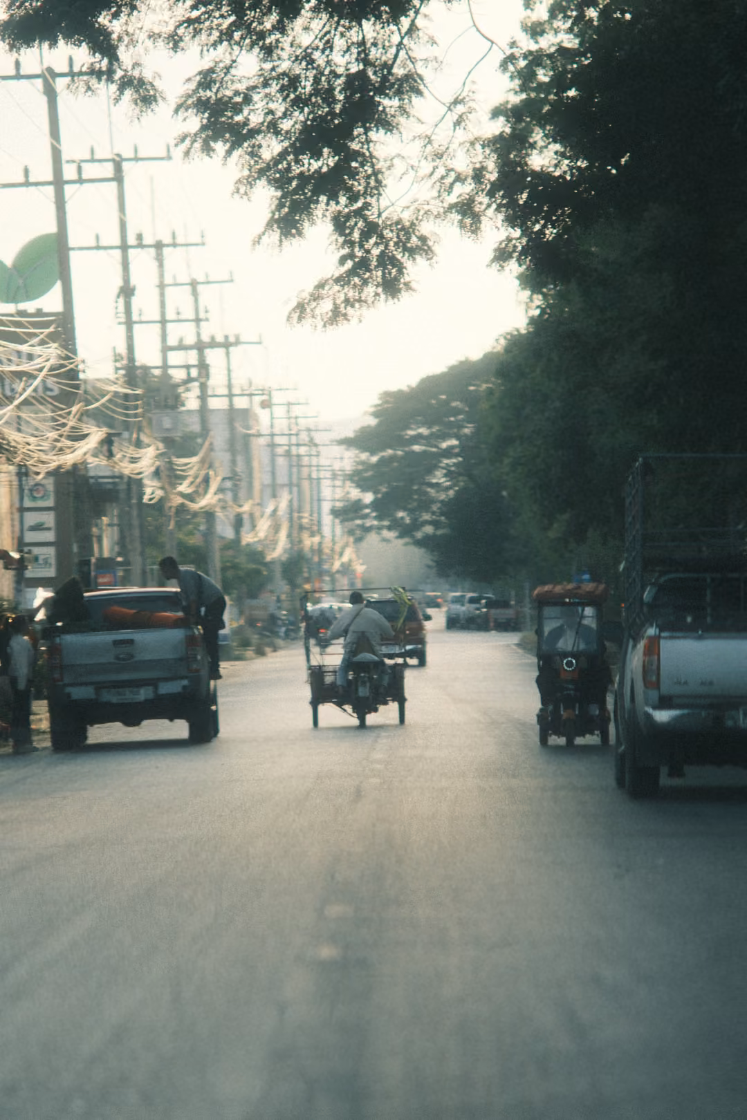 A hazy road seen from behind traffic: a pickup, a motorbike with a sidecar and a parked white pickup under trees.