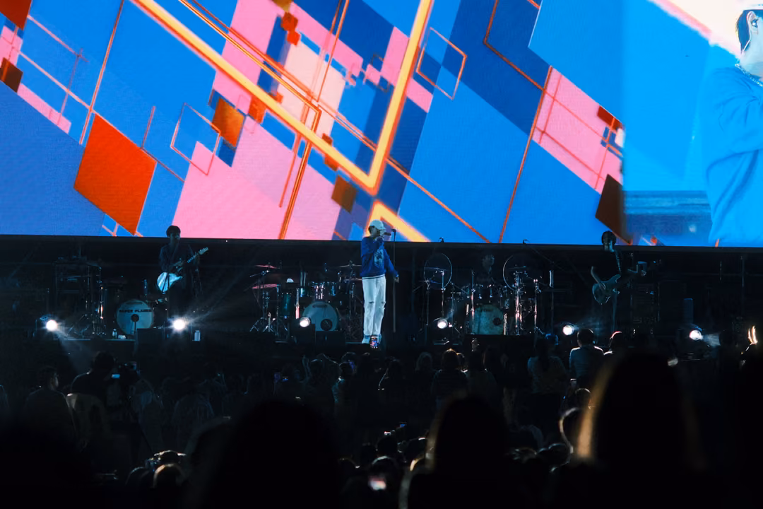 Band on stage below a screen of pink, orange and blue geometric diamonds, crowd silhouettes in front.