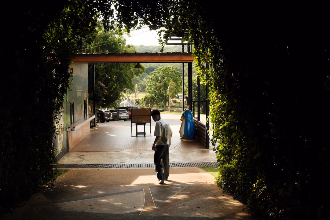 Silhouetted person walking through a dark leafy archway toward a bright hall with a car park beyond
