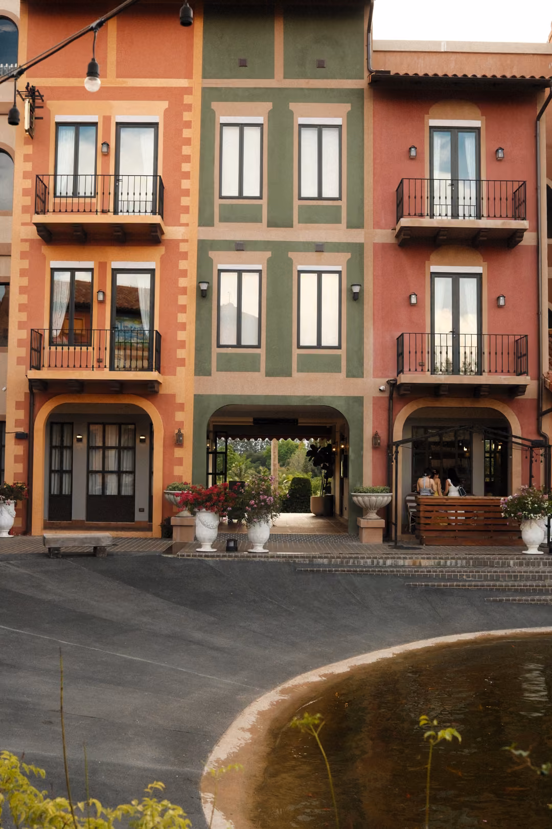 Orange, green and red facades with balconies facing a paved plaza and a round pond with reeds
