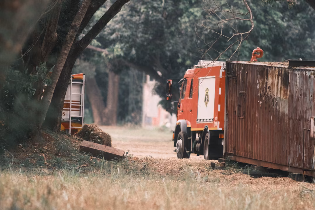 A red fire truck parked beside a rusted shipping container on a dirt track under large trees