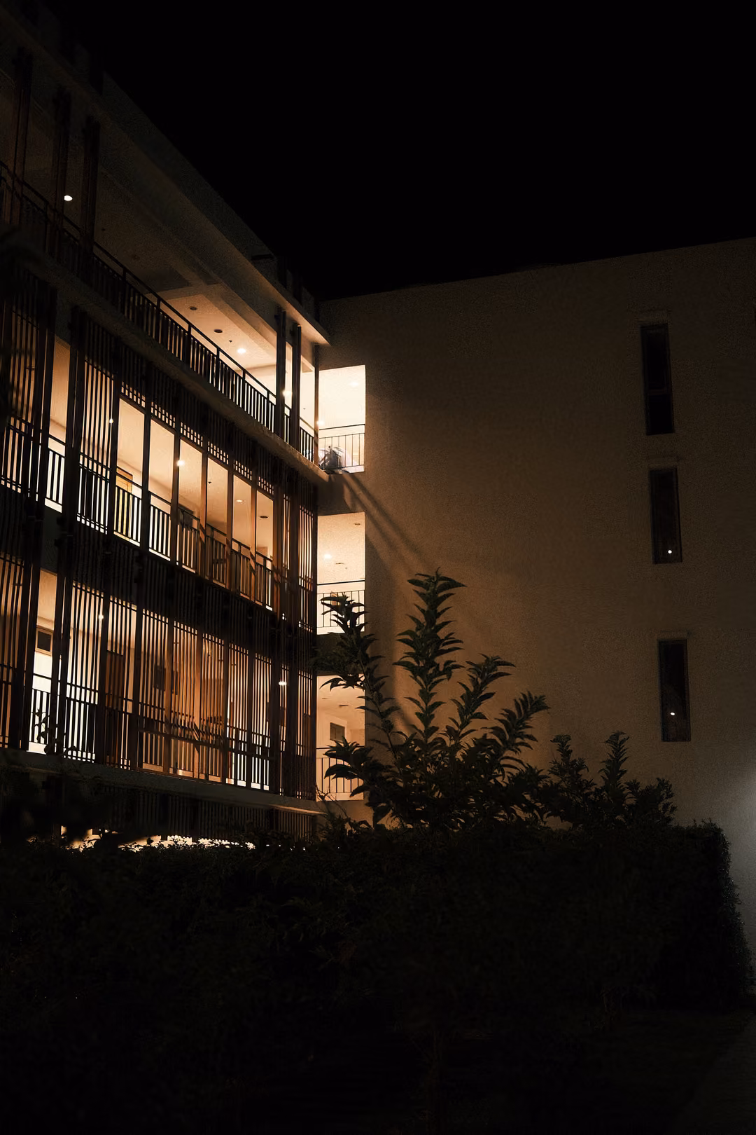 Side of a multi-storey building at night with warm lit balconies, a dark hedge and trees in front