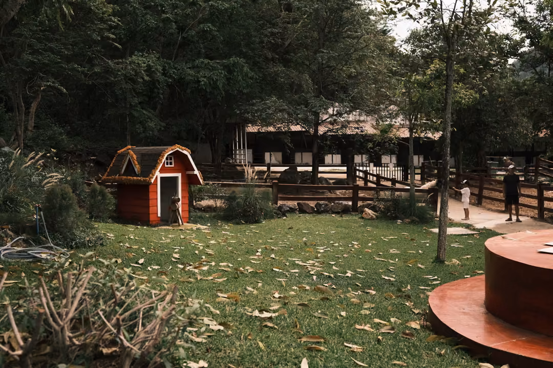 Red miniature barn on a leaf-scattered lawn ringed by wooden fences, trees and a red round platform