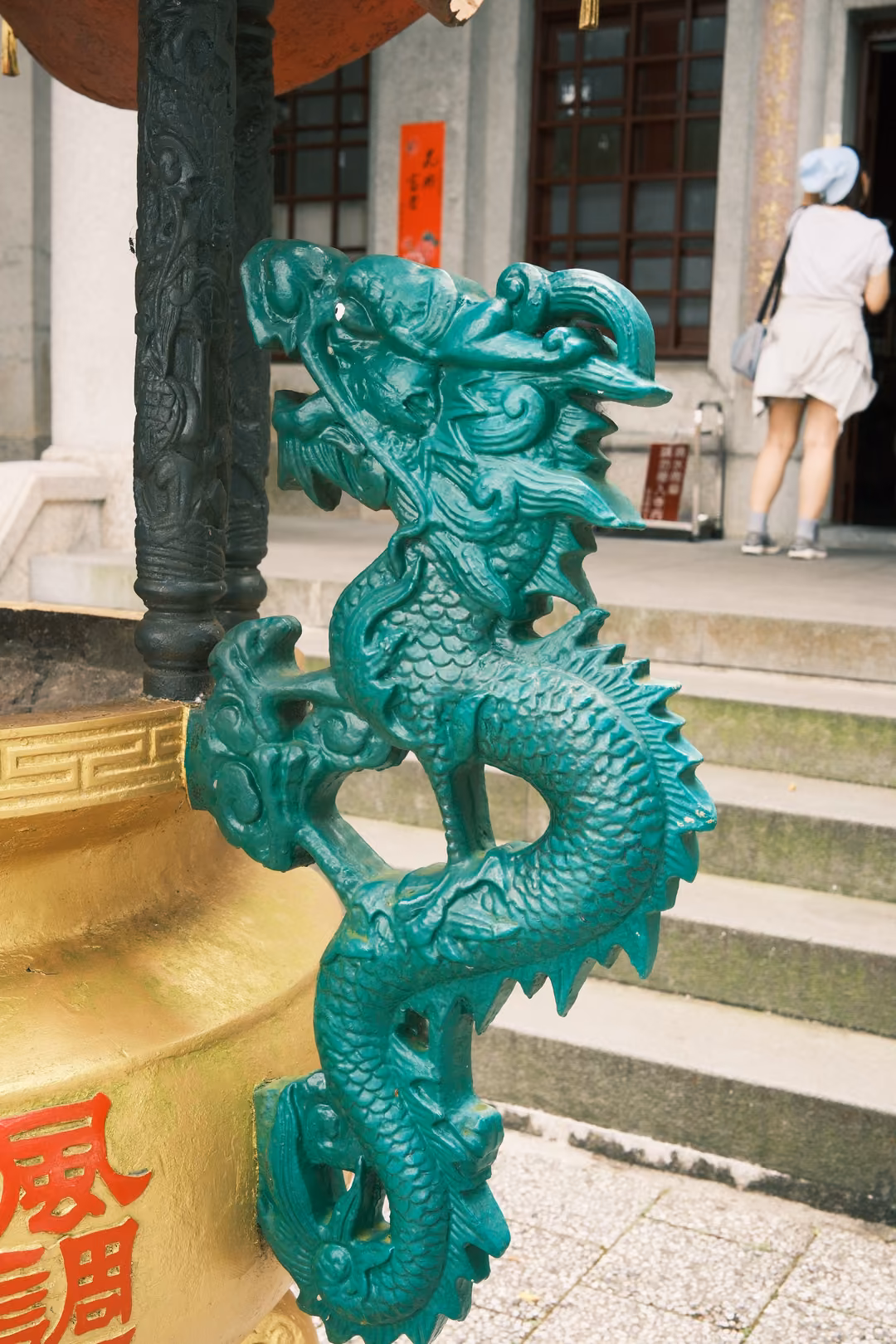 Green cast-metal dragon coiled on a golden urn beside temple steps, a visitor in white at a doorway