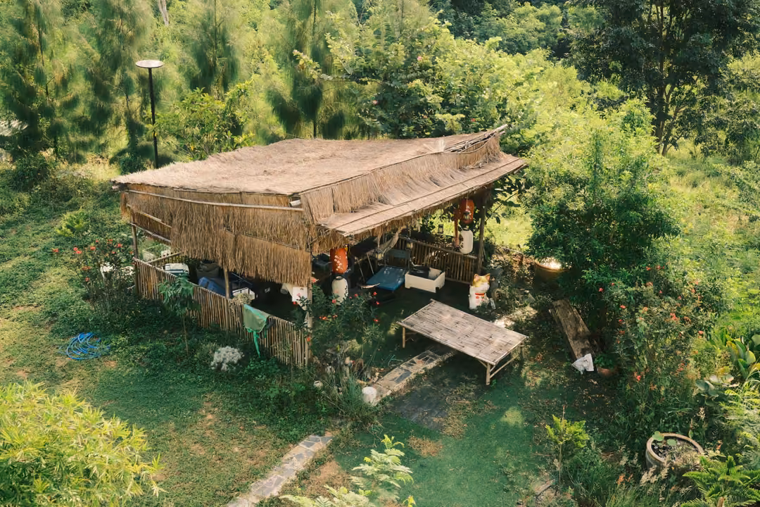 A thatched-roof hut seen from above among trees and grass, with a wooden bench and scattered items around it.