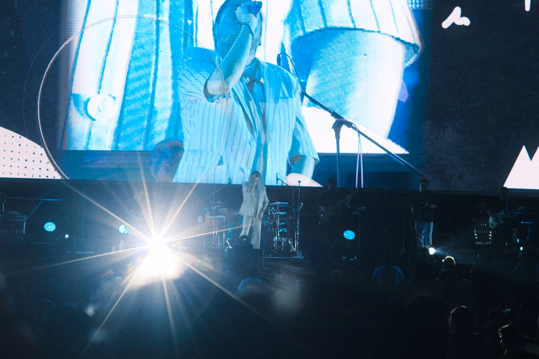 Angled view of the screen showing a raised arm and microphone stand, a strong starburst light flaring on the stage below