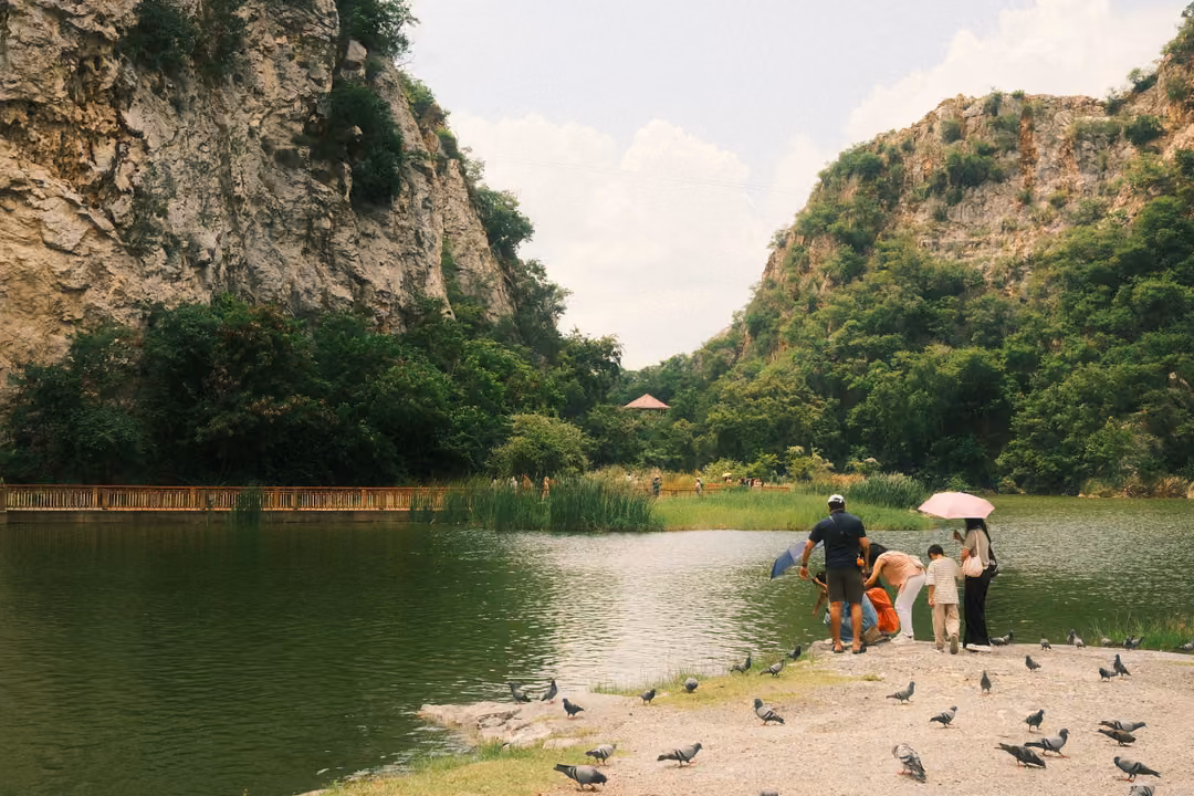 Visitors with an umbrella feeding pigeons on a lakeshore, cliffs and a cloudy blue sky behind