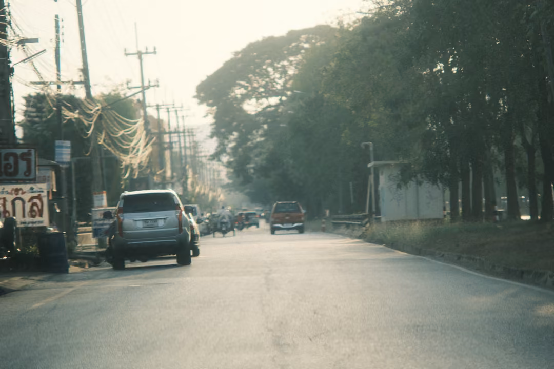 A silver SUV parked at the roadside under tall trees, a red Thai sign at left and cars fading into haze down the road.