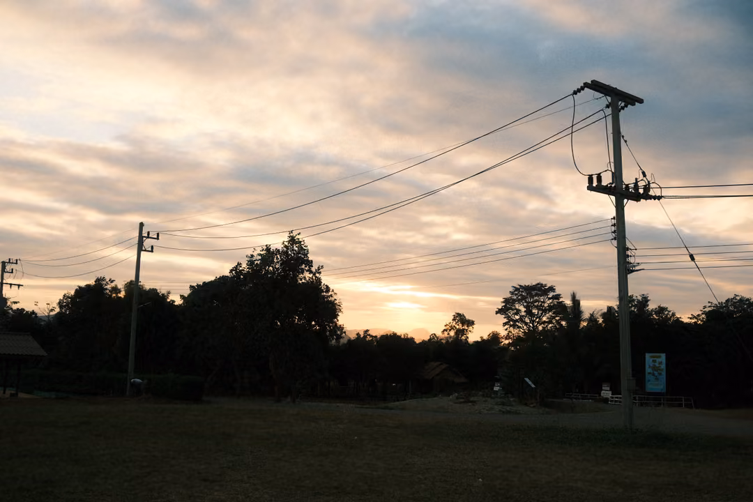 Silhouetted power poles and wires against a sunset sky with dark clouds, trees and an open field below