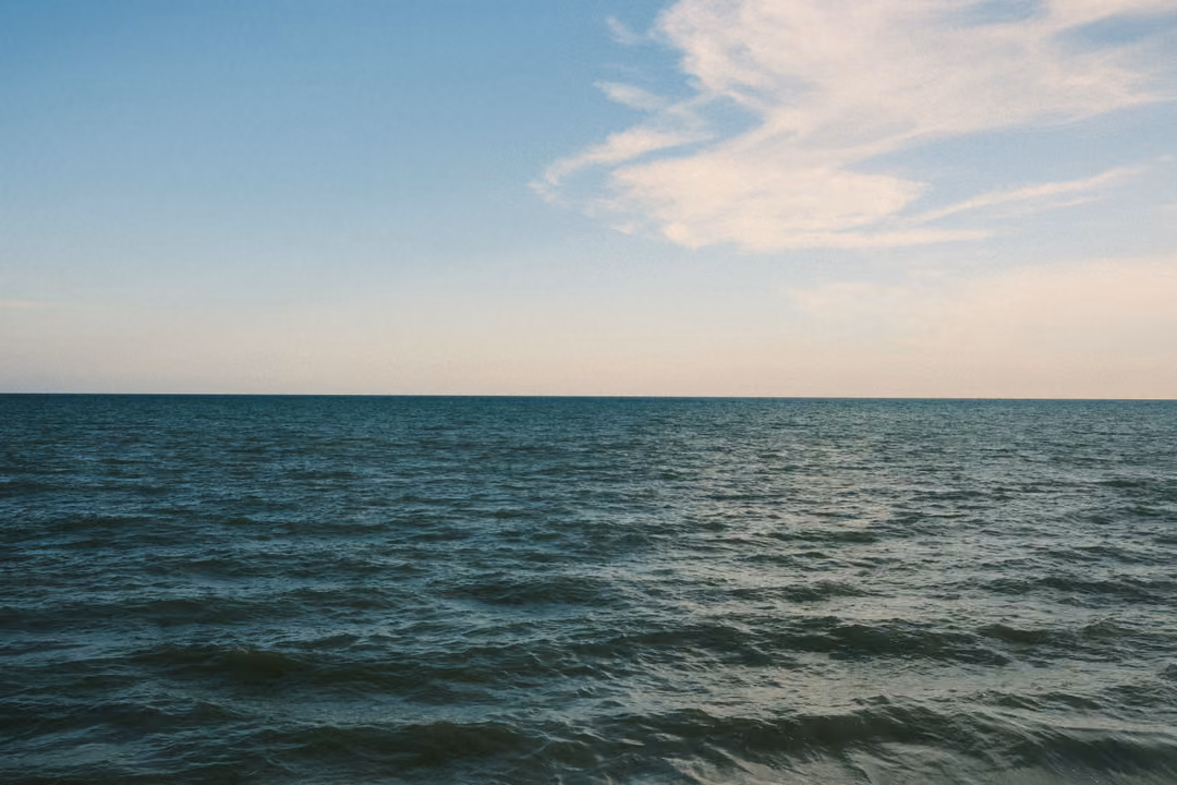 Open sea with gentle rolling waves under a pale blue sky with wispy clouds, no land visible