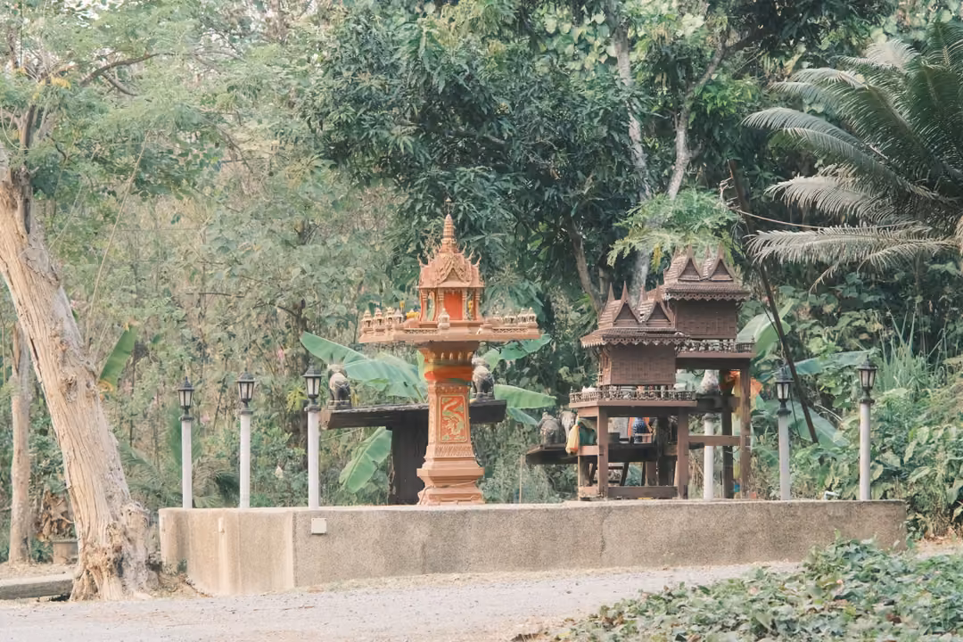 Ornate red and orange spirit house beside a wooden shrine on a concrete platform among trees