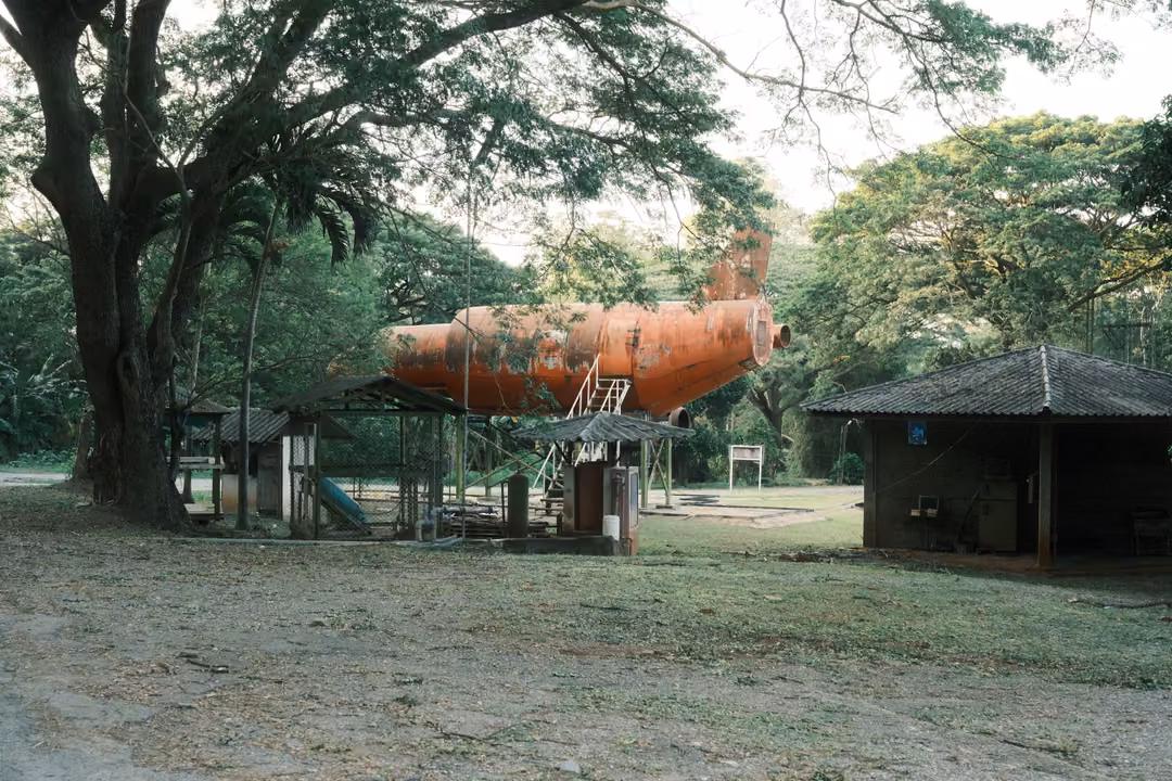 Rusted orange aircraft fuselage mounted on stilts under large trees, with a thatched shelter nearby