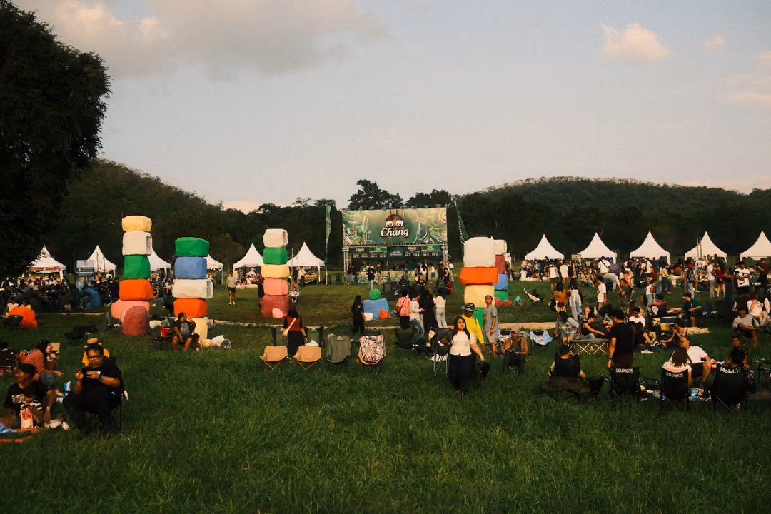 Stacked towers of colourful cubes rise from a crowded grassy field with white tents and a green banner.