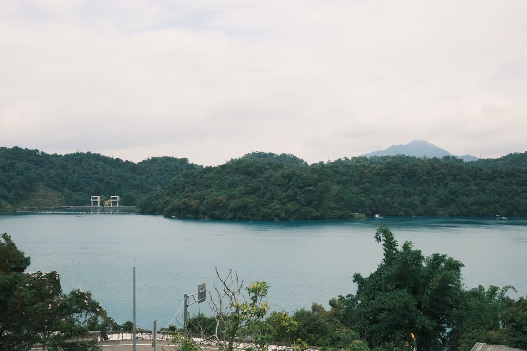 Broad lake panorama framed by treetops, a small structure on the far shore and a distant blue peak