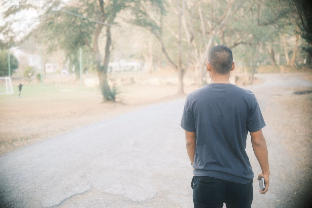 Man in a grey t-shirt seen from behind standing on a cracked road, trees and a distant field ahead