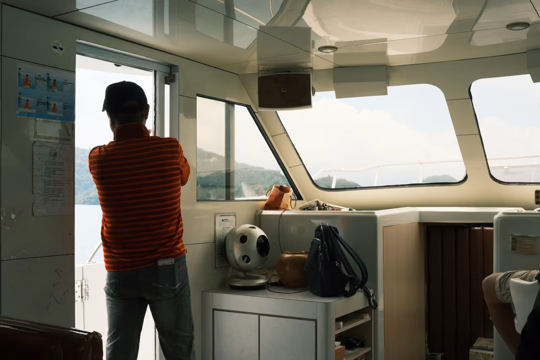 Man in a red striped shirt and cap seen from behind at a boat cabin doorway, windows showing the lake