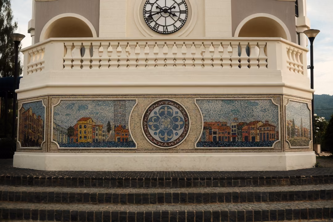 Base of a cream tower with a clock, a balustrade and mosaic panels showing colourful town buildings above steps