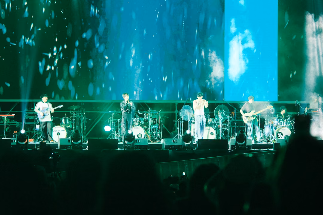 Stage with a screen of falling flakes and a cyan panel showing a blurred figure, band performing below.