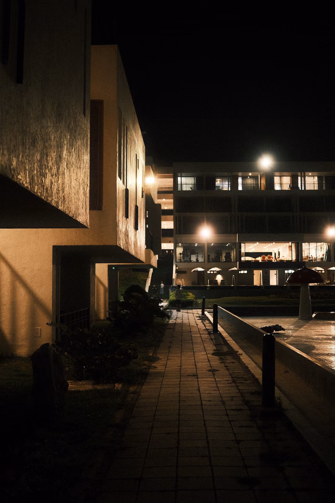 A tiled path between buildings at night leading toward a lit hotel block with umbrellas and street lamps