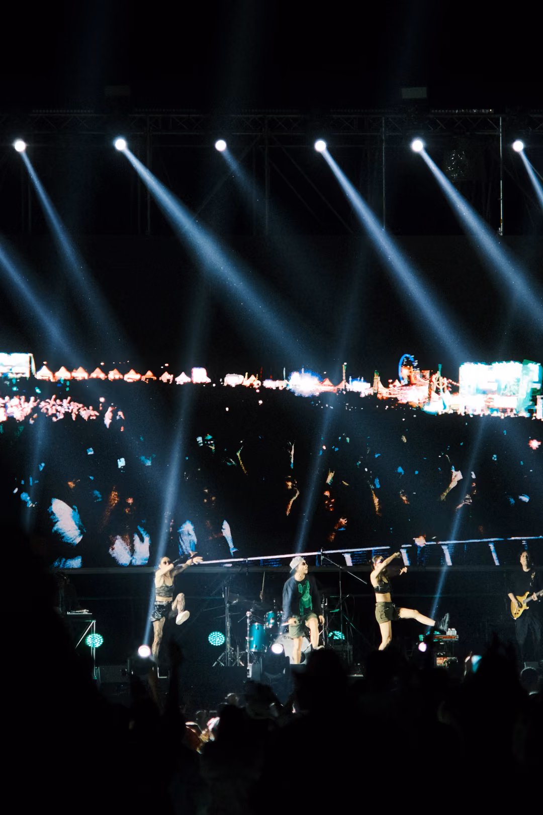 Vertical frame of white light beams angling down from the truss over performers and a screen showing the crowd