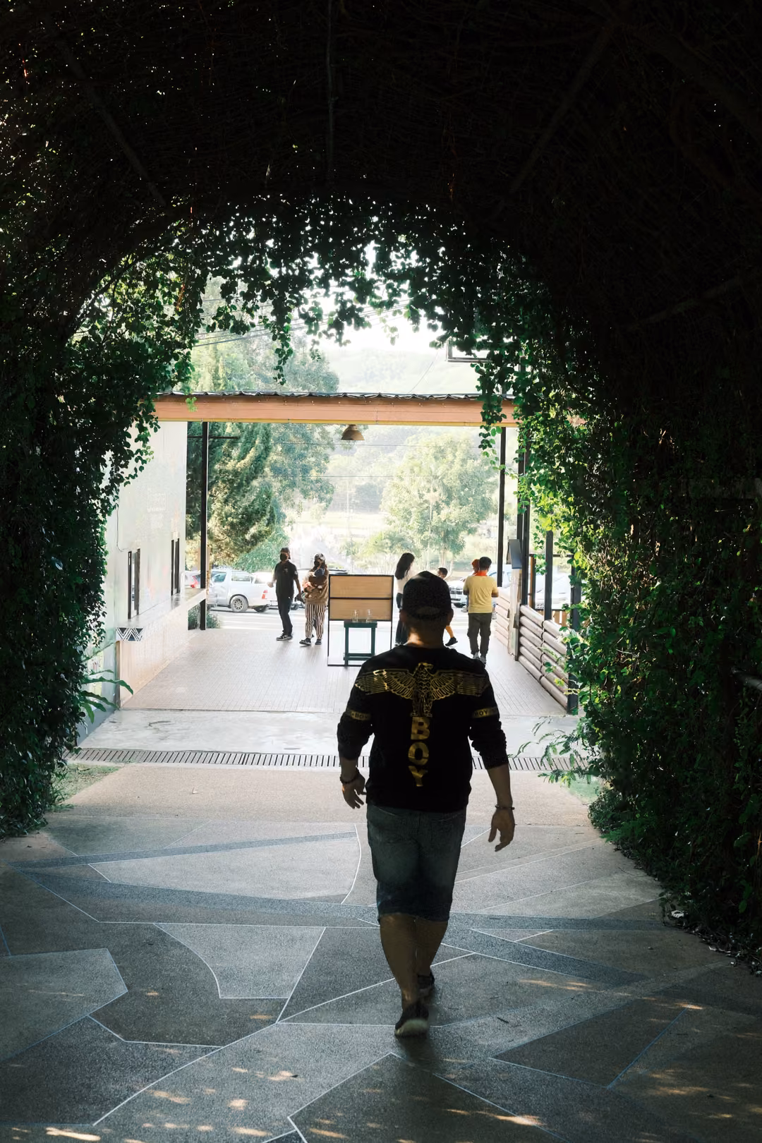 Man in a black shirt walking through a leafy archway tunnel toward a bright open entrance