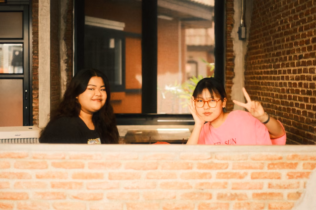 Two women seated behind a low brick wall, one in black smiling, one in pink with glasses making a peace sign