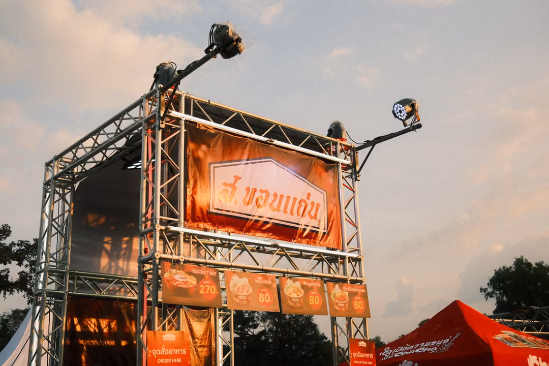 Steel truss tower with spotlights holding a red food-stall banner in Thai script under a pale late-afternoon sky.