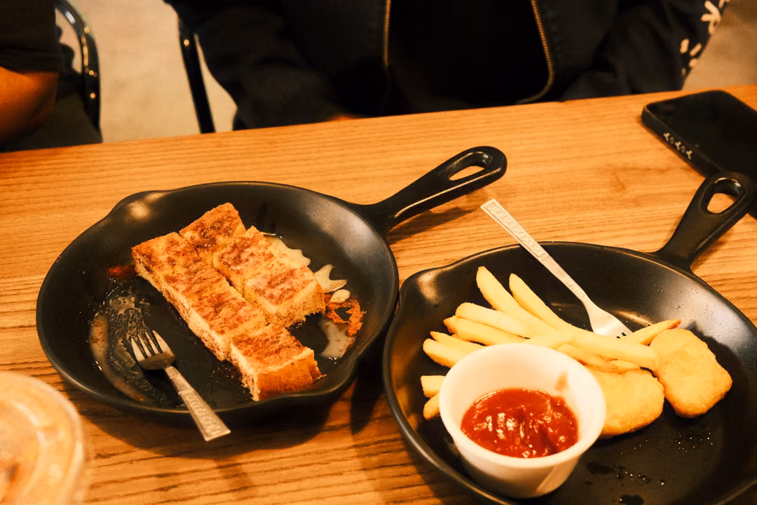 Wooden table with two black skillet pans: a slab of toast with a fork, and fries with nuggets and ketchup