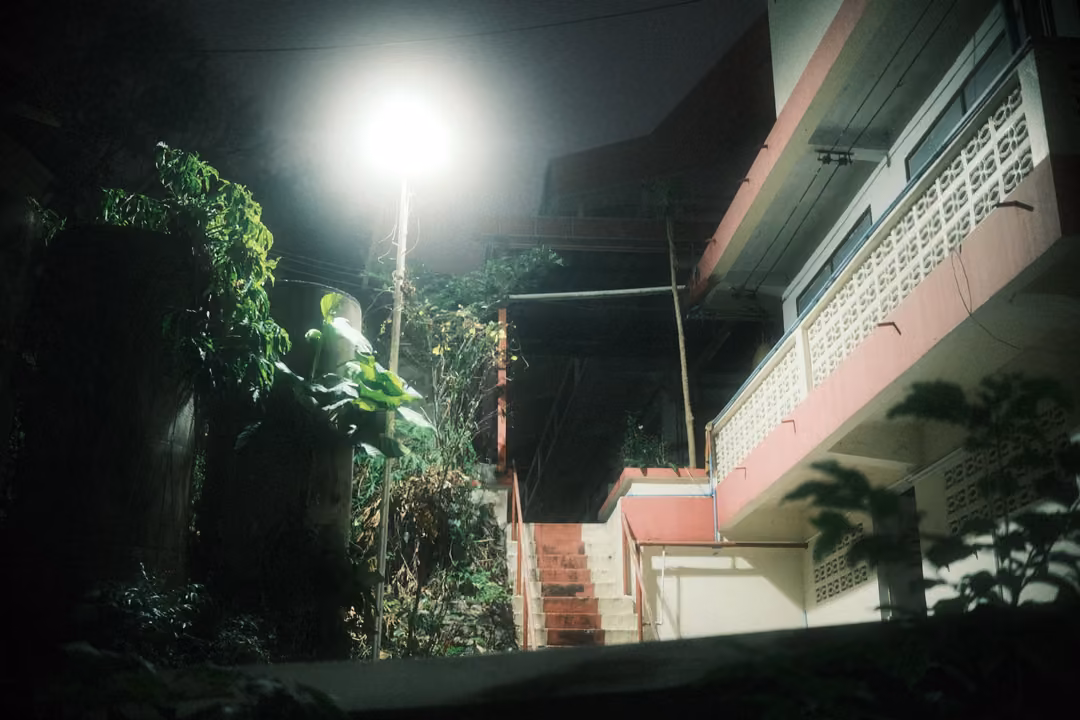 Building with a lattice balcony and outdoor stairs lit by a bright streetlamp at night, leafy plants