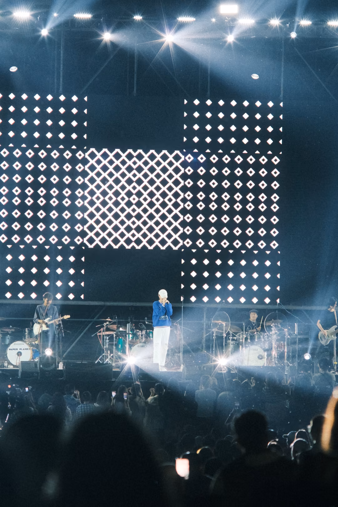 Vertical view of a screen of diamond grids above the singer standing in a white spotlight, crowd heads below.