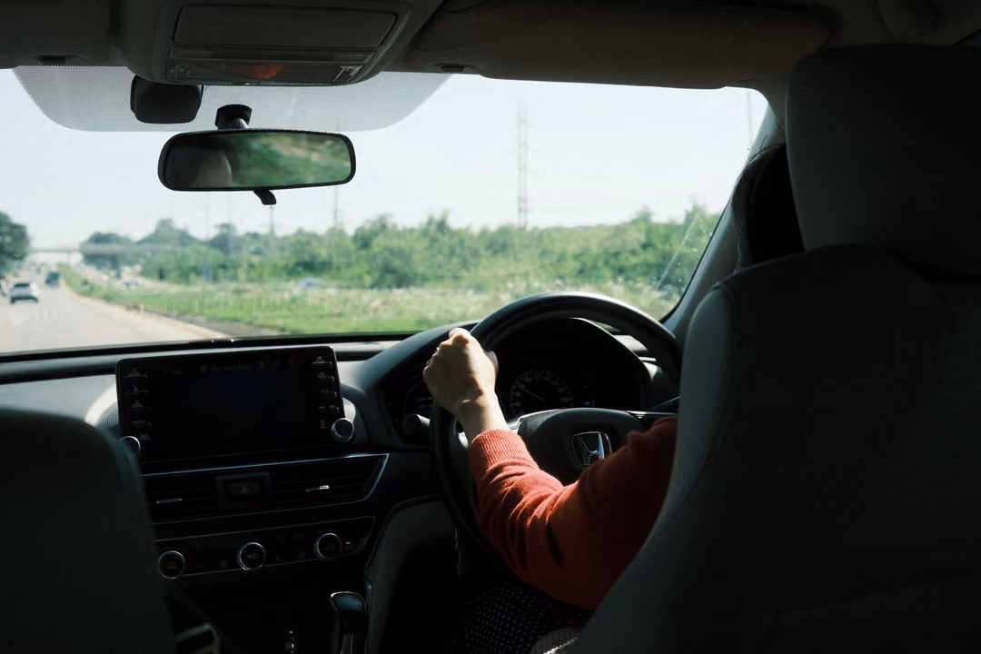 Car interior on a highway, a hand in a red sleeve on the wheel, open road and green verge ahead