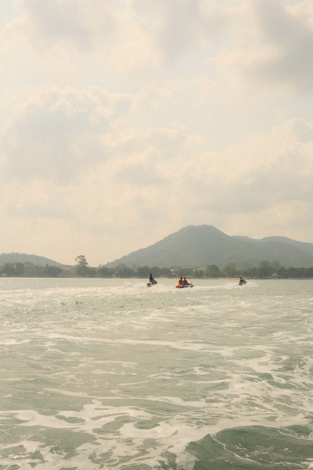 Three jet skis cross foamy water toward a green mountain under a hazy, cloudy sky.