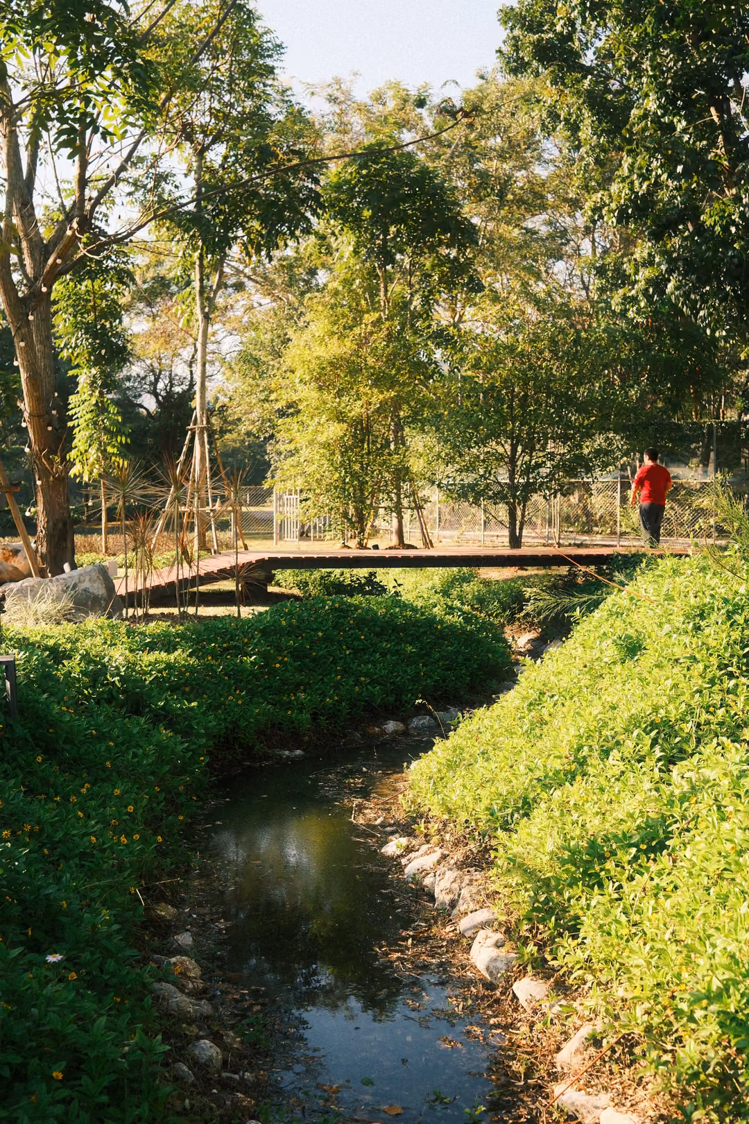 Stream between grassy banks leading to a footbridge, a person in a red shirt crossing the bridge