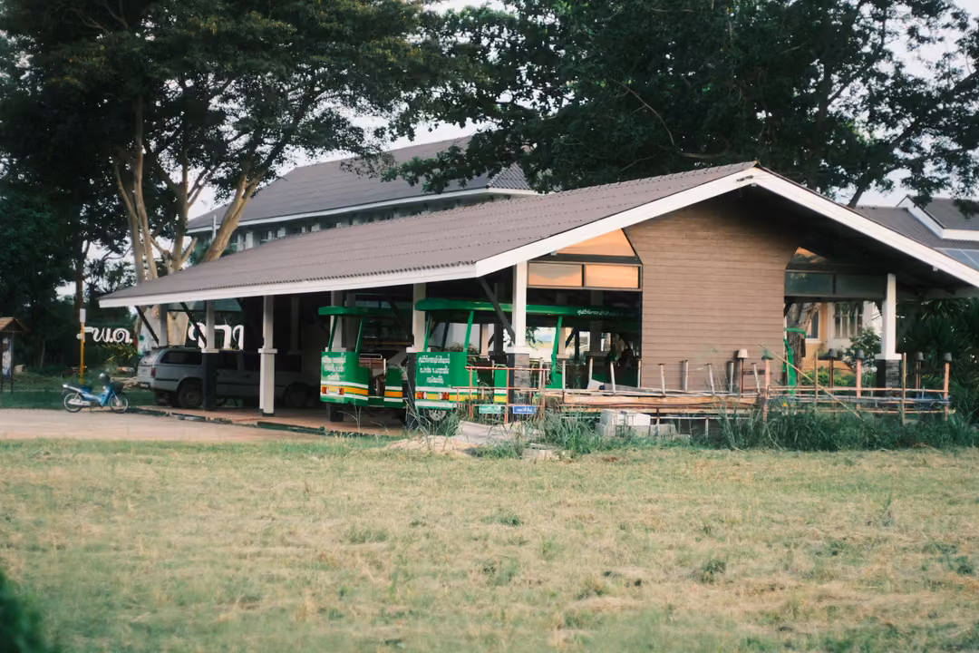 Shelter with a long sloped roof sheltering green open-air trams beside a field and trees