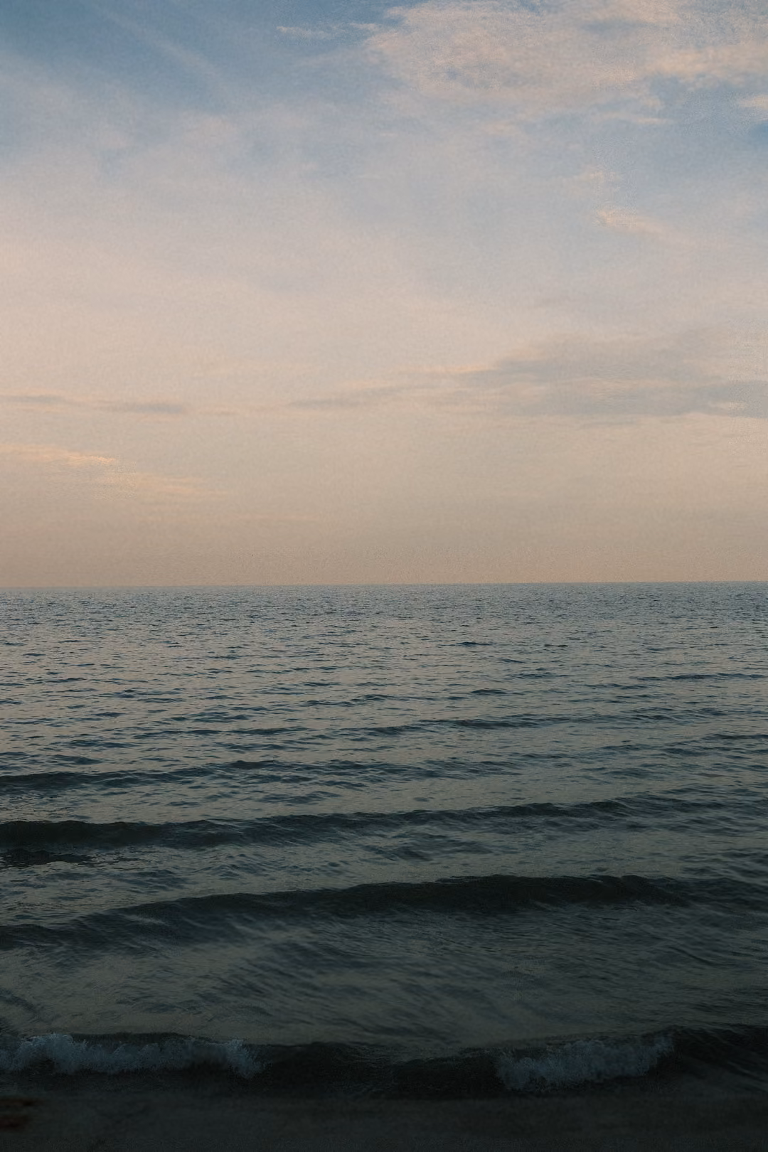 Vertical view of calm sea with small waves breaking near the shore under a pale, softly clouded evening sky