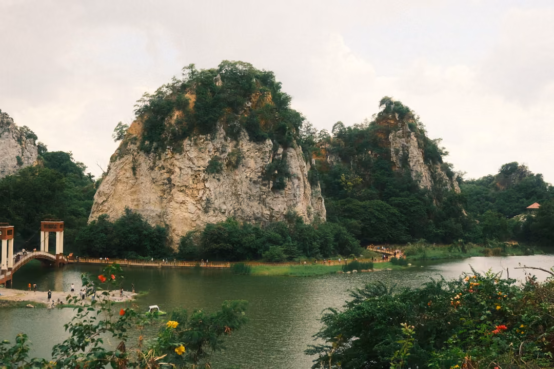 Elevated view of a large green-capped crag above a lake, bridge towers on the left, flowering shrubs in front