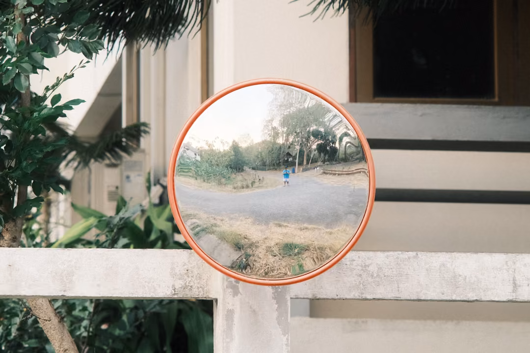 Round orange-framed convex mirror on a white wall reflecting a road, trees and a small figure